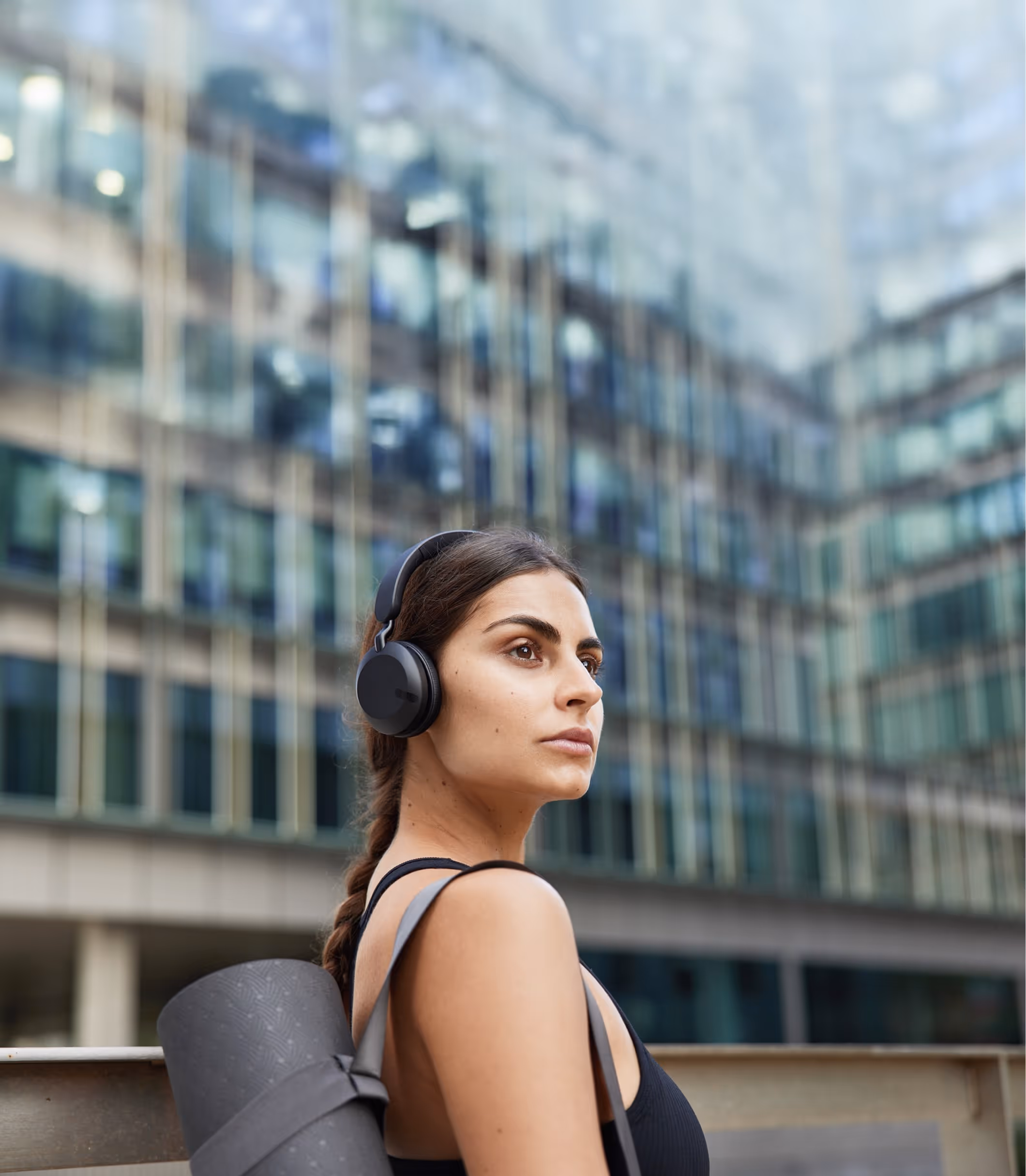 Young woman with braided hair wearing black headphones and a black tank top carrying a yoga mat outdoors in an urban setting.