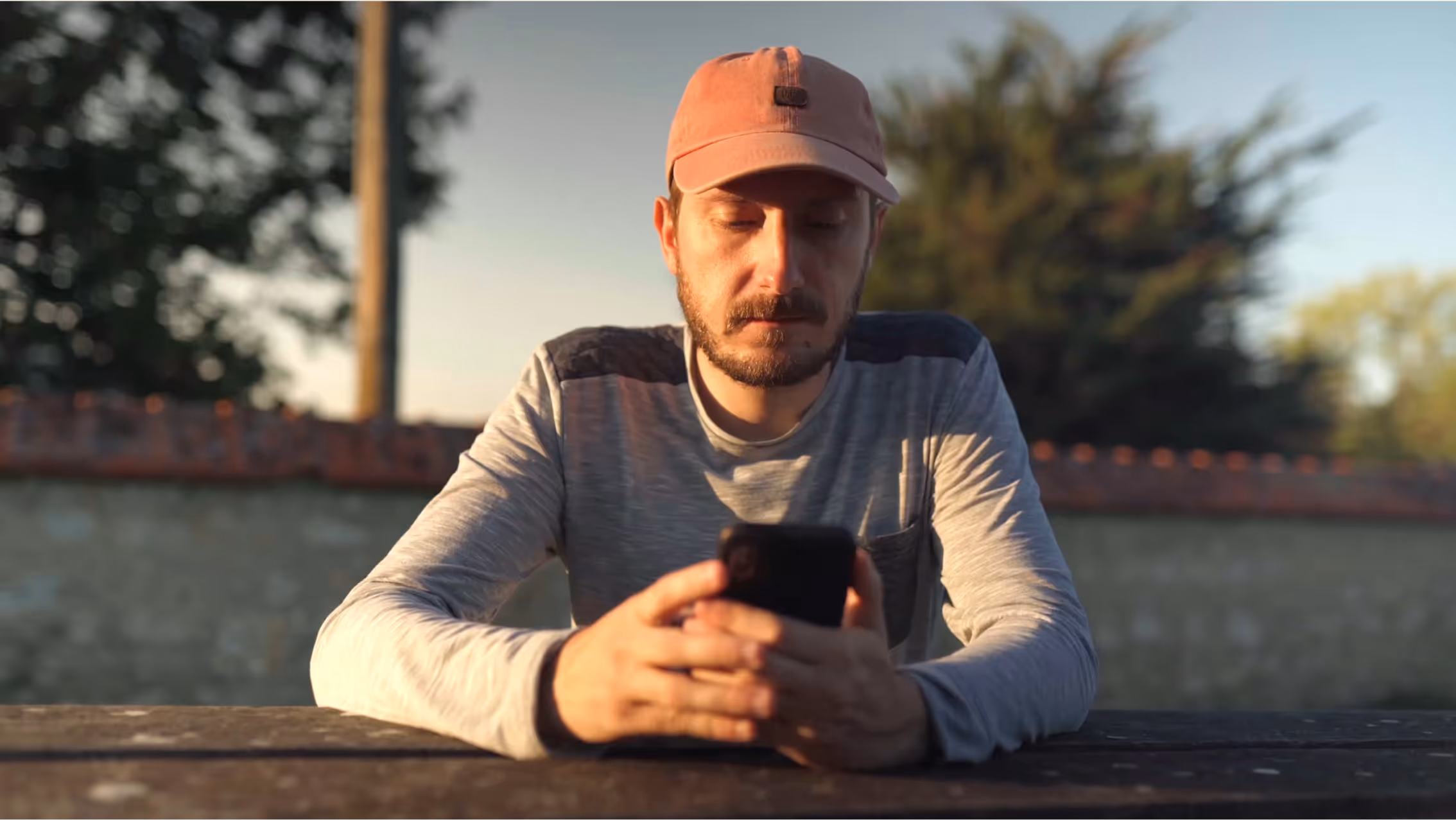 Man wearing a pink cap and gray shirt sitting at a wooden table outdoors, looking at his smartphone.